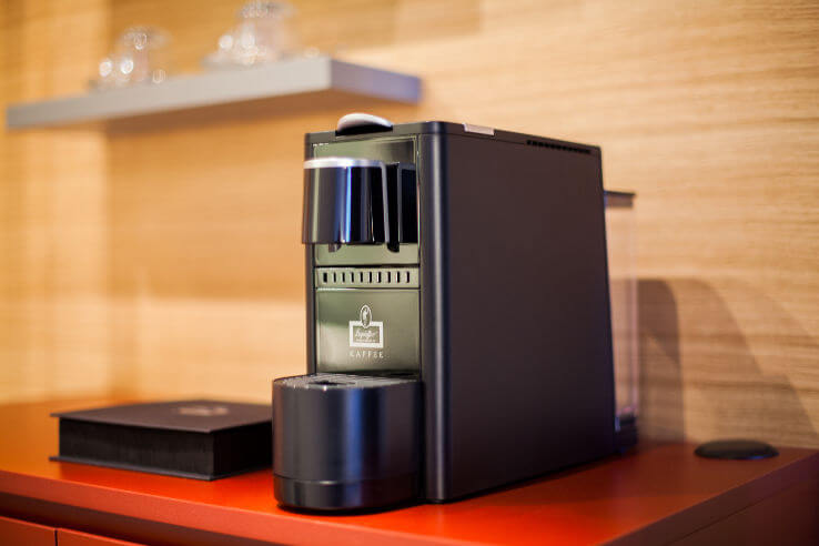 Leysieffer Kapselmaschine im Zimmer des ATLANTIC Hotel Aiport Kaffeemaschine auf rotem Tisch im ATLANTIC Hotel Airport Bremen, mit Gläsern im Hintergrund.