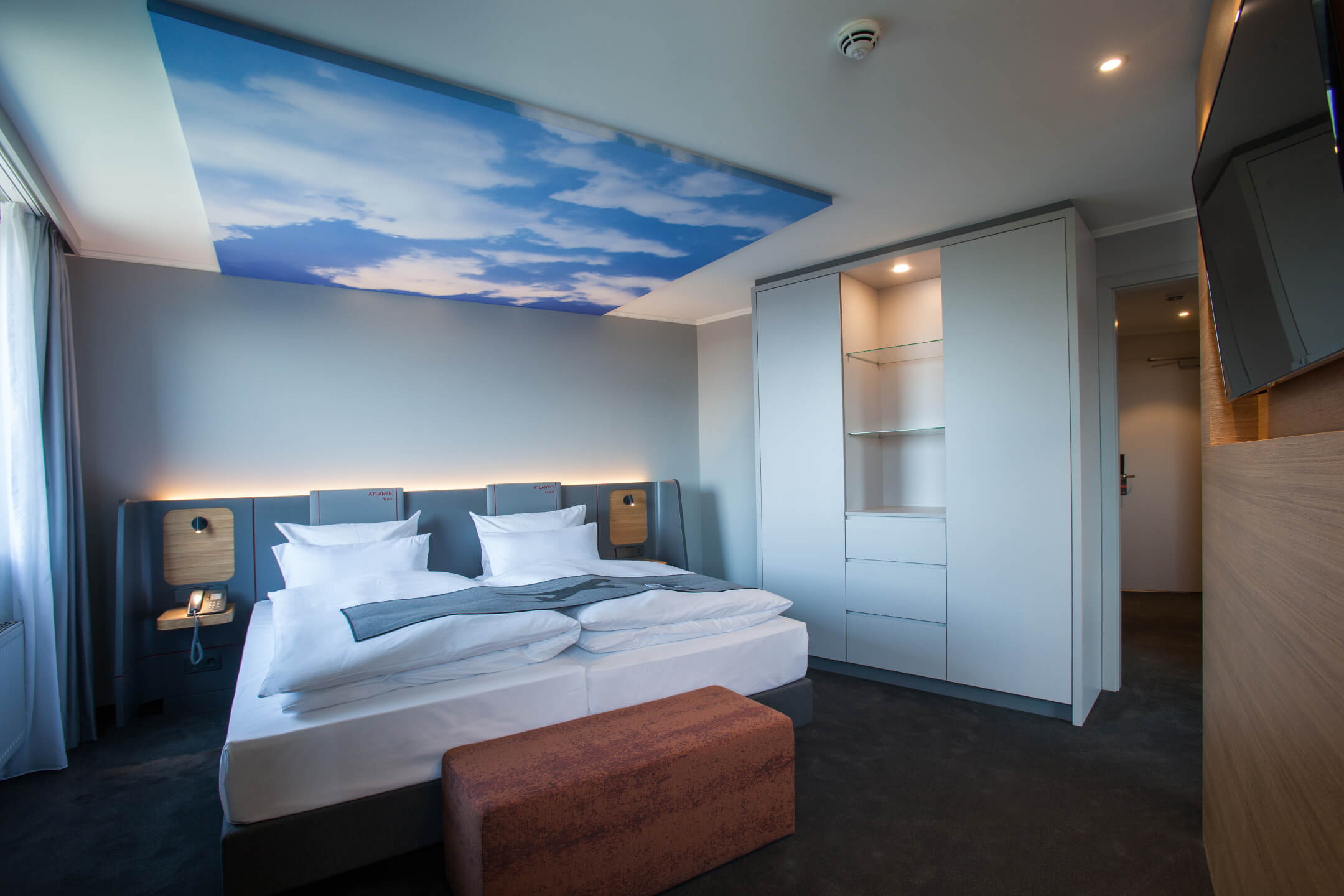 ATLANTIC Hotel Airport Bremen Suite Montgolfier Modernes Hotelzimmer mit Doppelbett, Himmelmotiv an der Decke, Schrank und Sitzbank im ATLANTIC Hotel Airport Bremen.