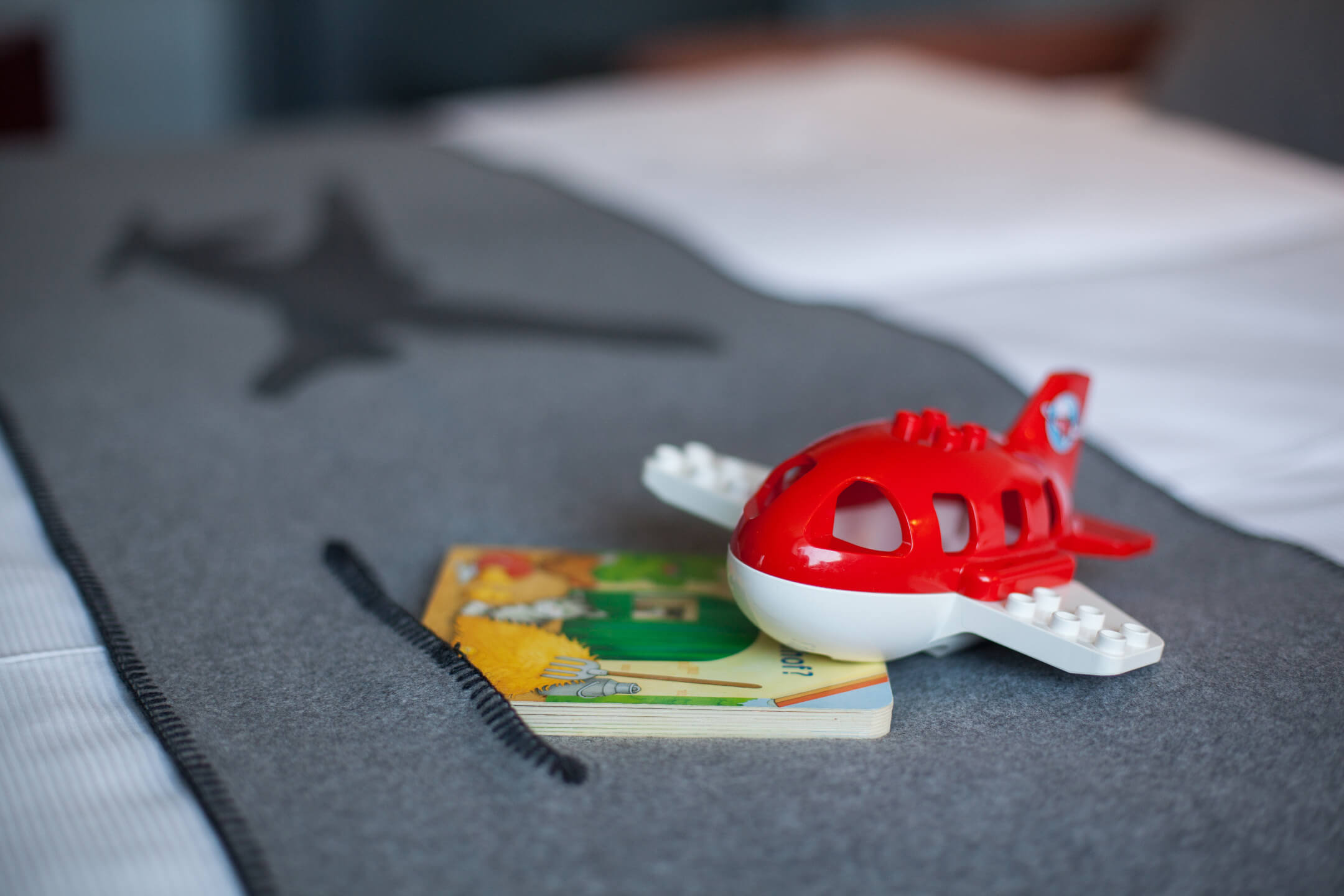 ATLANTIC Hotel Airport Bremen Kinderspielzeug in der Suite Montgolfier Rotes Spielzeugflugzeug und Buch auf grauer Decke im ATLANTIC Hotel Airport Bremen.
