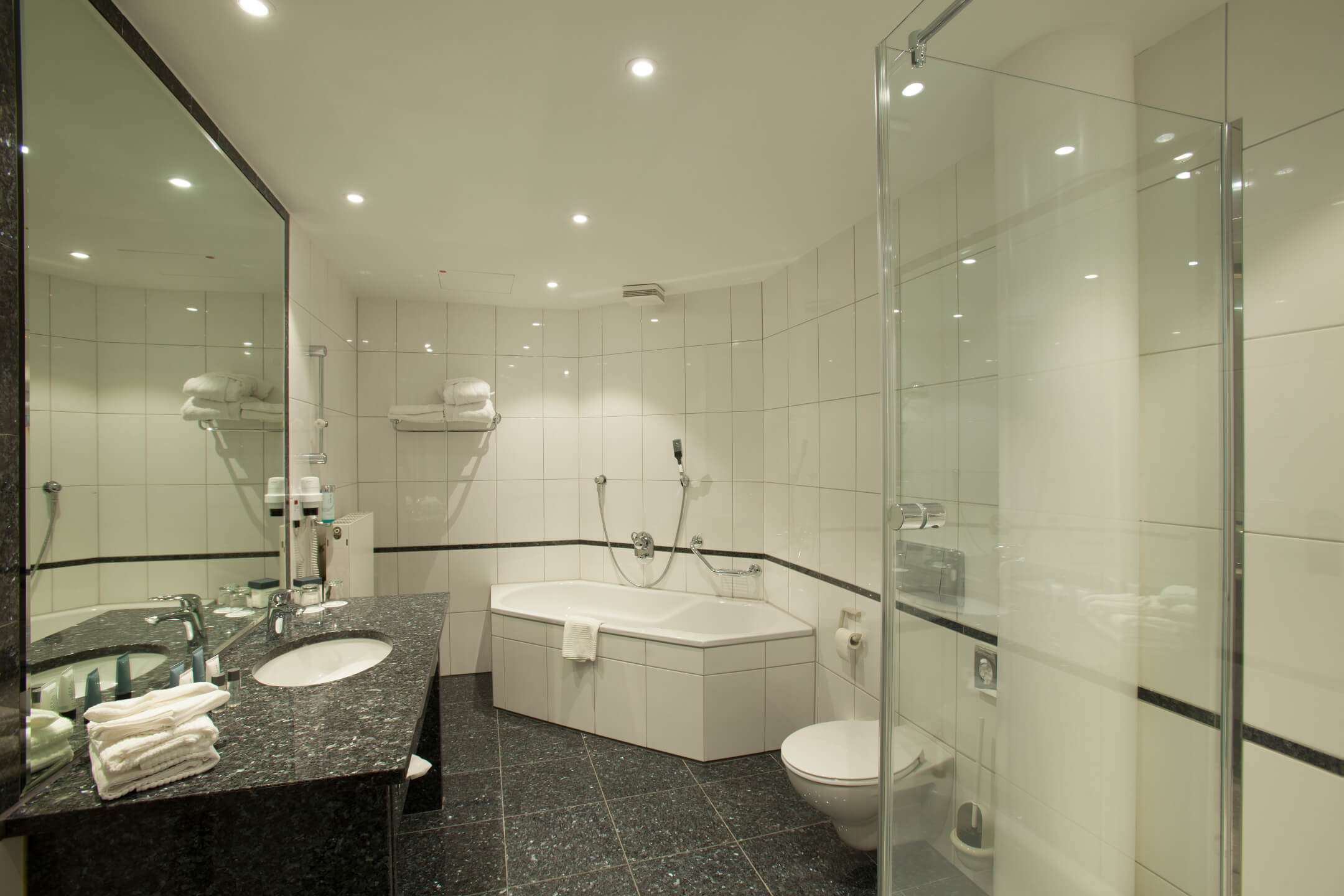 ATLANTIC Hotel Airport Bremen Badezimmer Suite Modernes Badezimmer mit großer Badewanne, Dusche, Doppelwaschbecken und weißen Fliesen im ATLANTIC Hotel Airport Bremen.