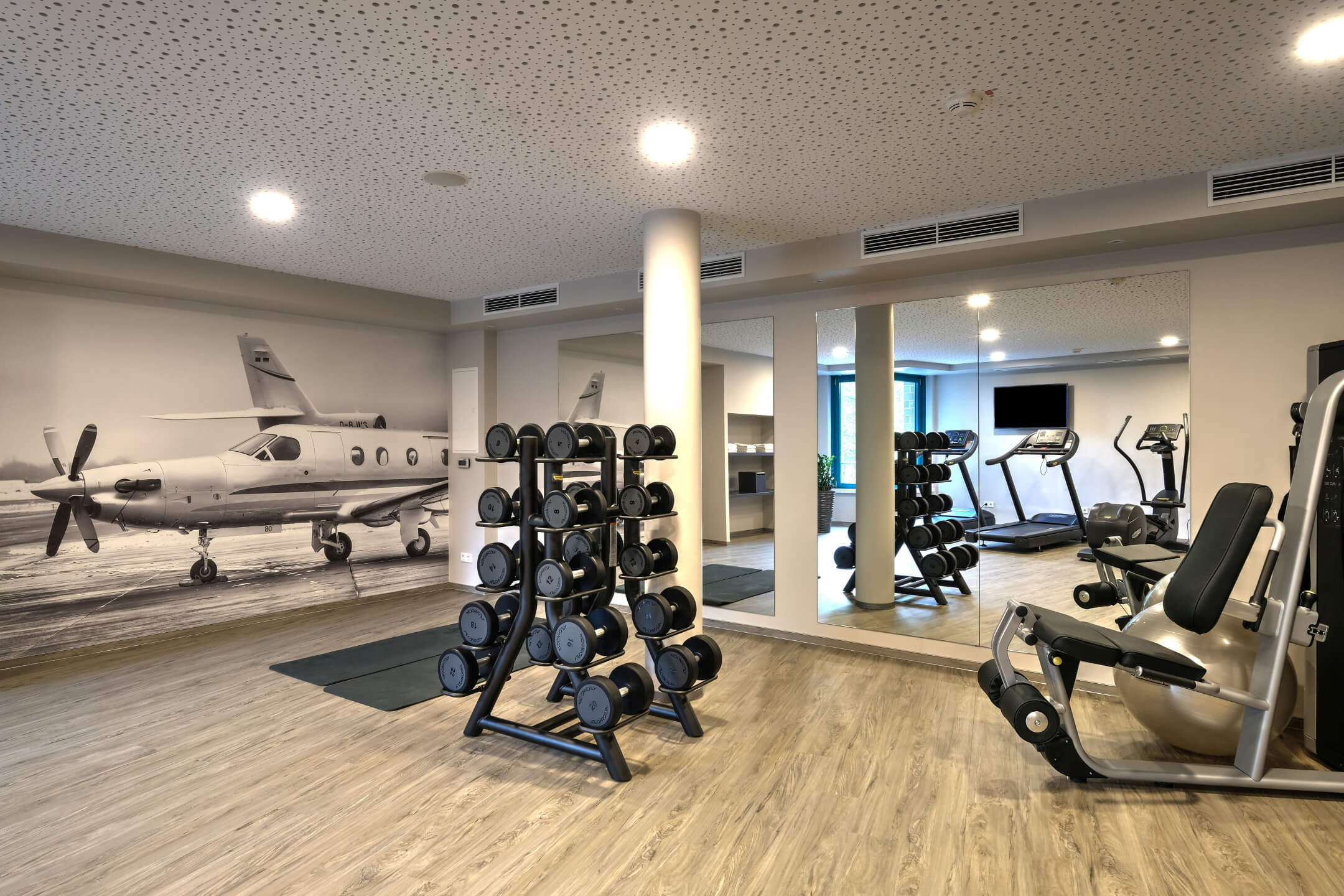 Fitness Raum in the ATLANTIC Hotel Airport Bremen Fitnessraum im ATLANTIC Hotel Airport Bremen mit Hanteln, Fitnessgeräten und Flugzeugbild an der Wand.