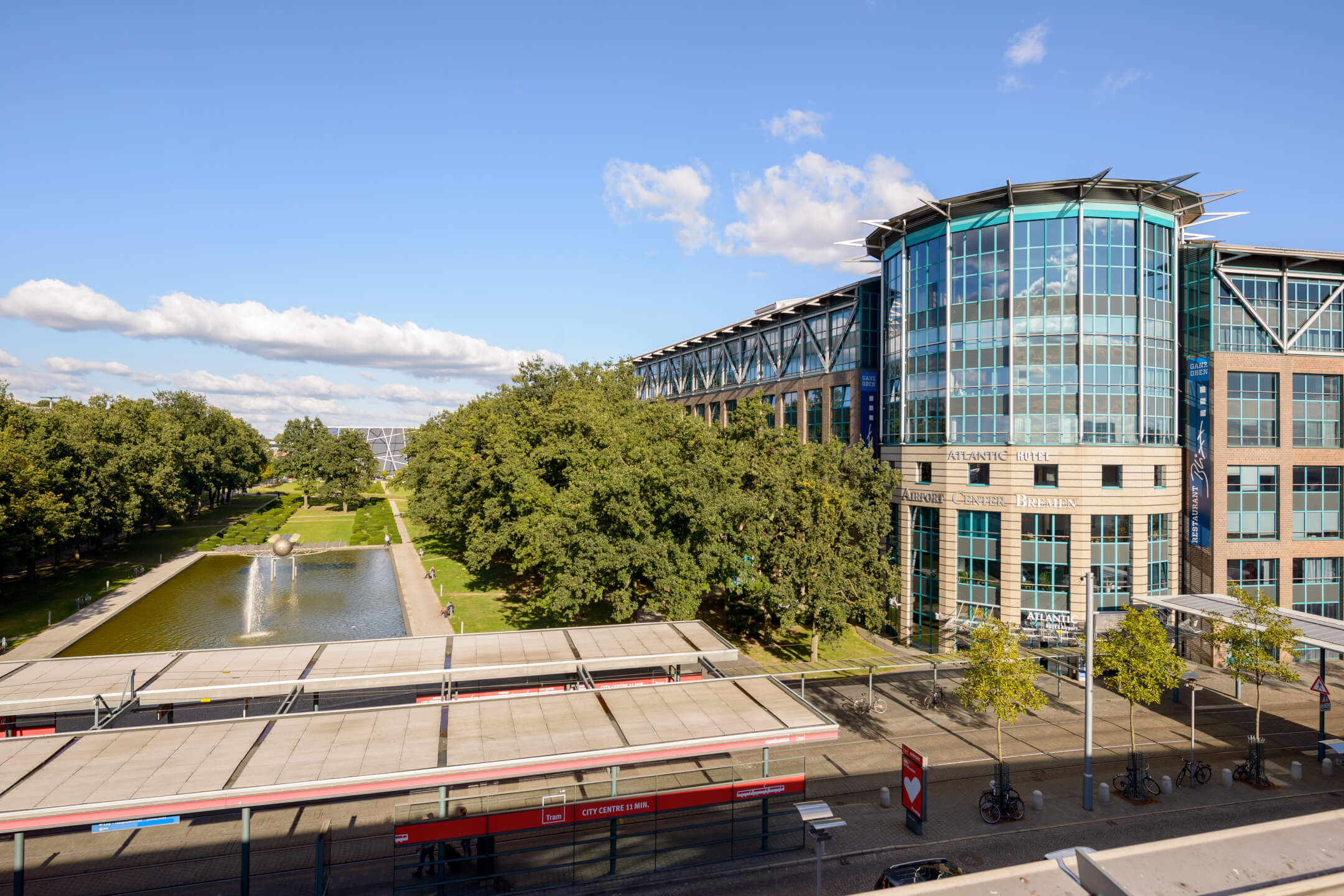 ATLANTIC Hotel Airport Bremen mit moderner Glasfassade, Park mit Teich und Bäumen im Vordergrund, blauer Himmel.