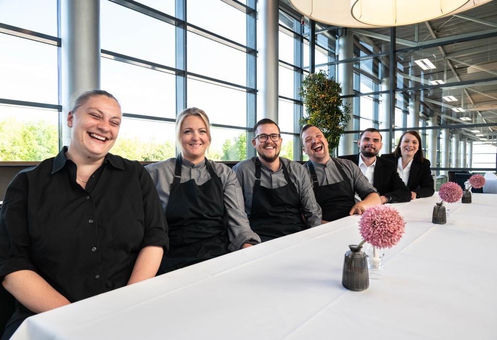 Bestens betreut im Gottlieb Restaurant Bremen Hotelteam sitzt lachend an einem Tisch mit weißen Tischdecken und rosa Blumen, große Fenster im Hintergrund.
