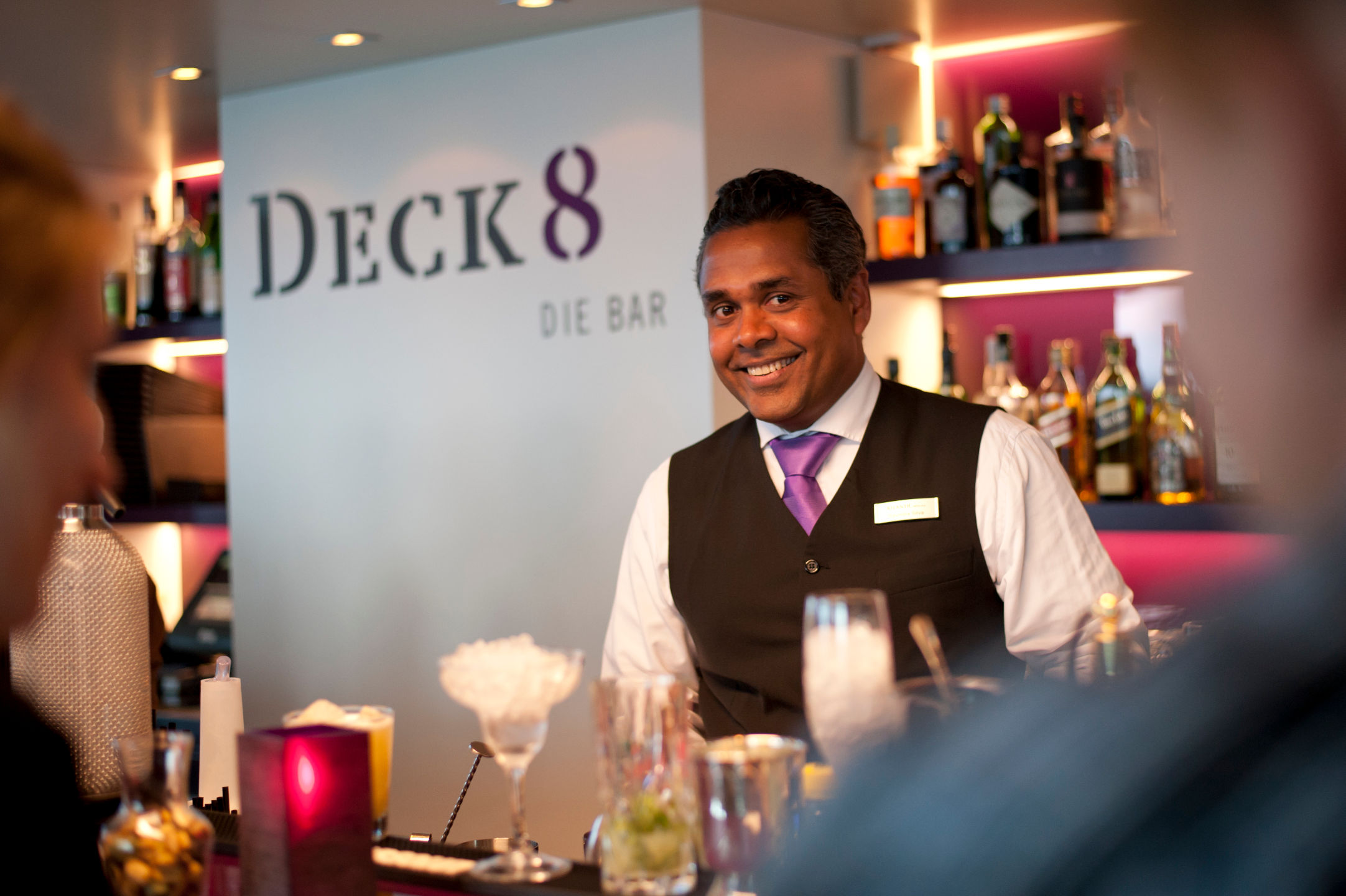 ATLANTIC Hotel Kiel – Barchef Barkeeper in eleganter Weste lächelt hinter der Theke der stilvollen Bar "Deck 8", umgeben von Flaschen und Gläsern.