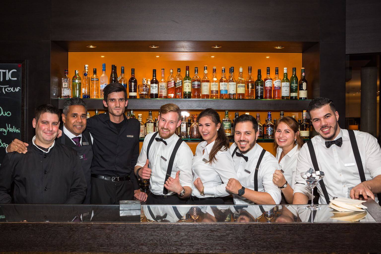 Barevent im ATLANTIC Grand Hotel Bremen Barteam posiert lächelnd vor einer gut bestückten Bar, alle in eleganter Uniform.