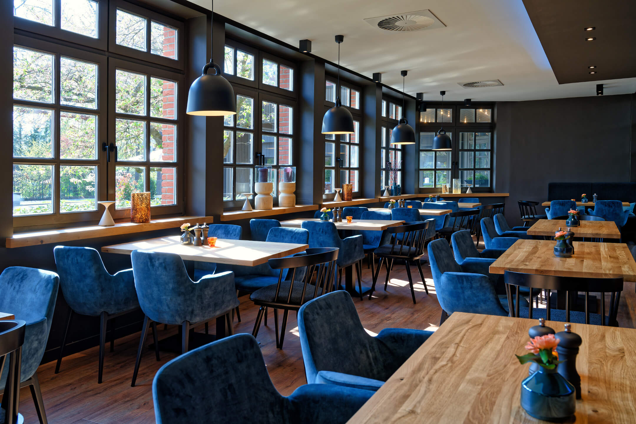 Restaurant VICTORIAN im ATLANTIC Hotel Landgut Horn Helles Restaurant mit großen Fenstern, Holztischen, blauen Polsterstühlen und moderner Beleuchtung im ATLANTIC Hotel.