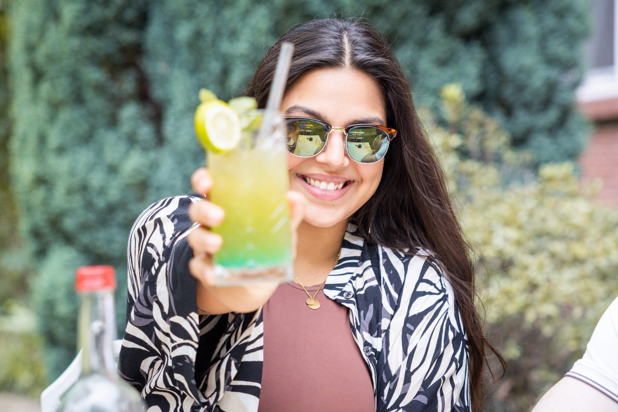 Drinks im ATLANTIC Hotel Landgut Horn Bremen Frau mit Sonnenbrille hält lächelnd einen Cocktail in der Hand, im Garten des ATLANTIC Hotel Landgut Horn Bremen.