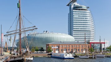 Außenansicht auf das Gebäude des ATLANTIC Hotel SAIL City Bremerhaven im Hintergrund Hafenansicht mit Segelschiff, modernem Glasgebäude und historischem Backsteingebäude bei klarem Himmel.