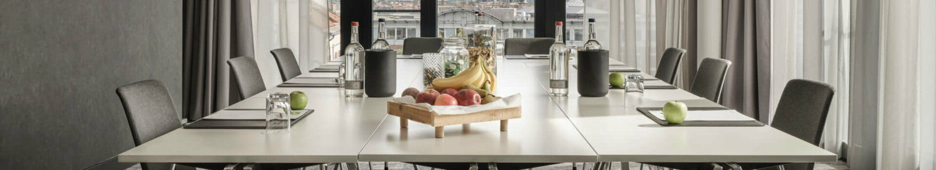 ATLANTIC Hotel Münster Tagungsangebot 15 % Moderner Konferenzraum im ATLANTIC Hotel Münster mit großem Tisch, Stühlen und einem Obsttablett, lichtdurchflutet durch Fenster.