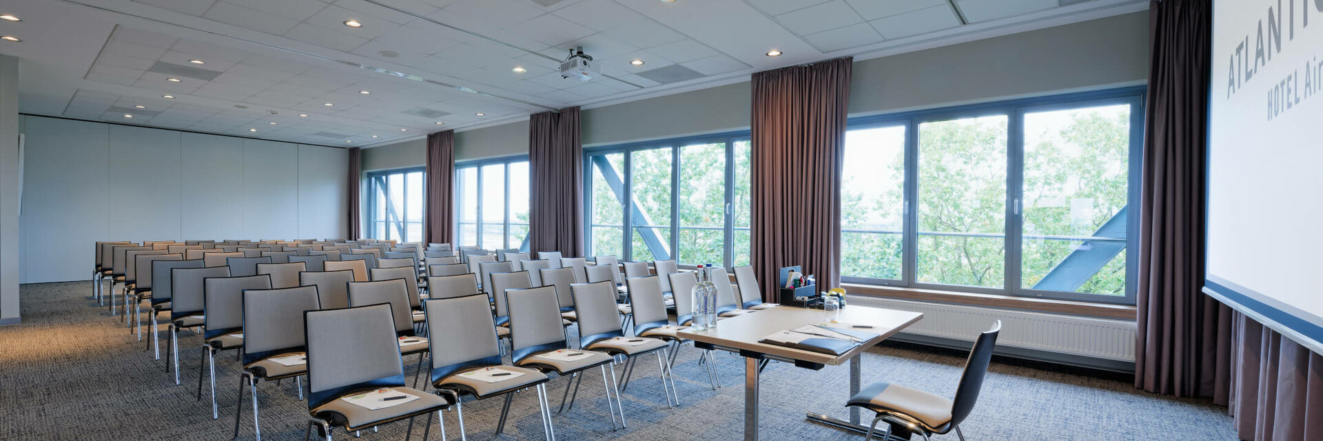 ATLANTIC Hotel Airport Tagungsraum Heller Konferenzraum im ATLANTIC Hotel Airport Bremen mit Stuhlreihen, Tisch und großen Fenstern mit Blick ins Grüne.
