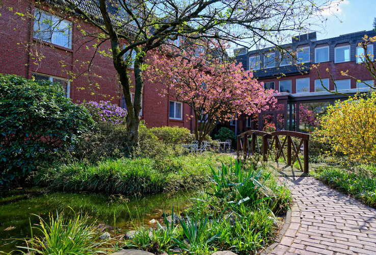 Garten im ATLANTIC Hotel Landgut Horn Bremen Gartenansicht mit blühenden Bäumen, kleinem Teich, Holzbrücke und rotem Backsteingebäude im ATLANTIC Hotel Landgut Horn.