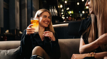 ATLANTIC Hotel Heidelberg | Lachende Frauen mit Drink in der Bar 15 High Zwei lachende Frauen in einer stilvollen Hotelbar, eine hält ein Getränk.