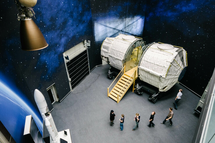 Space Lab | Raumfahrt-Führung Bremen Raumfahrt-Ausstellung mit Raketenmodell, Raumkapsel-Nachbildung und Besuchern vor einem Weltraum-Hintergrund.