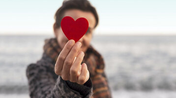 Gutschein-Shop ATLANTIC Hotels Person hält ein rotes Herz vor unscharfem Meereshintergrund.