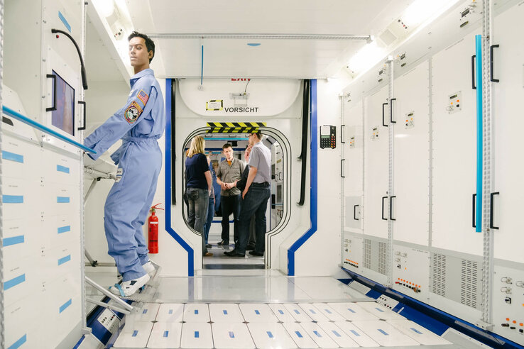 Columbus Modul | Raumfahrt-Führung Bremen Person in blauem Raumanzug in einem weißen, futuristischen Korridor mit Technik, im Hintergrund stehen mehrere Personen.