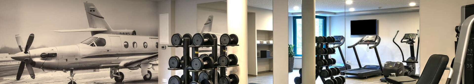 Fitnessbereich ATLANTIC Hotel Airport Bremen Fitnessraum im ATLANTIC Hotel Airport Bremen mit Hanteln, Fitnessgeräten und Flugzeugbild an der Wand.