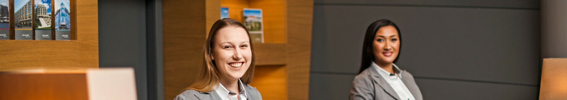 Rezeption im ATLANTIC Hotel Airport in Bremen Zwei Empfangsmitarbeiterinnen in grauen Anzügen lächeln an der Rezeption des ATLANTIC Hotel Airport Bremen.