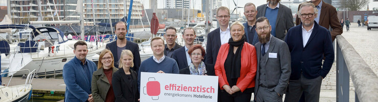 Energiekonsens Hotellerie Gruppe von Personen posiert mit einem Schild am Hafen, Boote und moderne Gebäude im Hintergrund.