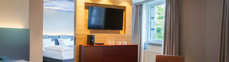 ATLANTIC Hotel Airport Bremen Suite Piccard Modernes Hotelzimmer mit Sitzbereich, Schreibtisch, TV, großem Fenster und Blick auf das Schlafzimmer im ATLANTIC Hotel Airport.