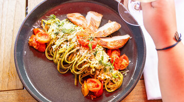 Essen im ATLANTIC Hotel Landgut Horn Bremen Teller mit Pasta, gegrilltem Hähnchen, Kirschtomaten und Kräutern, Hand hält ein Glas Wein.