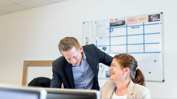 Ansprechpartner ATLANTIC Hotel Wilhelmshaven Zwei Personen in Business-Kleidung lachen gemeinsam in einem Büro vor einem Whiteboard.