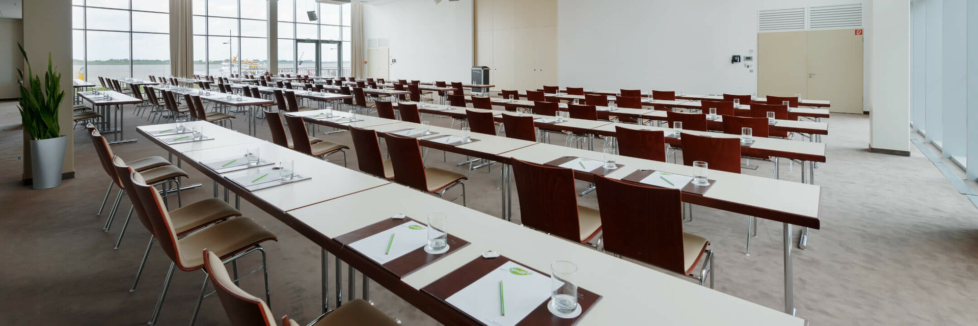 Saal Conference im ATLANTIC Hotel Sail City Moderner, heller Konferenzraum mit Reihen aus Tischen und Stühlen, großen Fenstern und Schreibmaterialien.