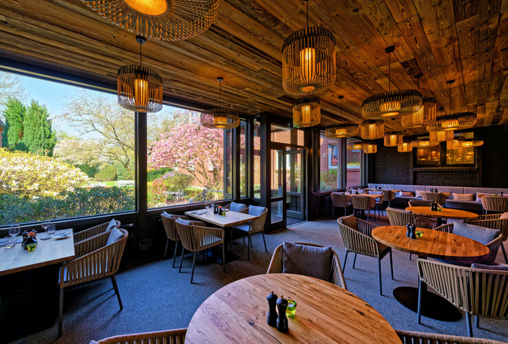 Wintergarten im ATLANTIC Hotel Landgut Horn Gemütliches Restaurant mit Holztischen und -decken, großen Fenstern mit Gartenblick und stilvollen Lampen.