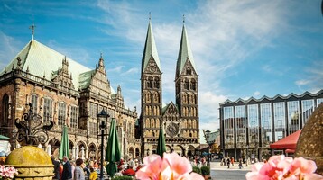 Historisches Gebäude und Kirche mit Doppeltürmen auf belebtem Platz bei blauem Himmel.