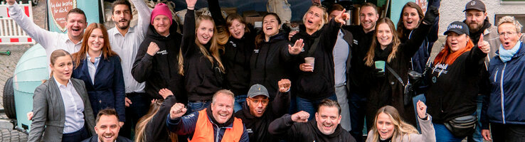 Gruppe fröhlicher Menschen in Freizeitkleidung posiert draußen vor einem bunt geschmückten Anhänger.