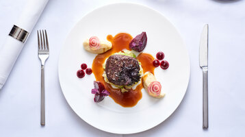 Foto-Banner für das ATLANTIC Grand Hotel Bremen auf der Aktionsseite Gourmetgericht mit Fleisch, Soße, Gemüse und roten Beeren auf weißem Teller, elegant gedeckter Tisch.