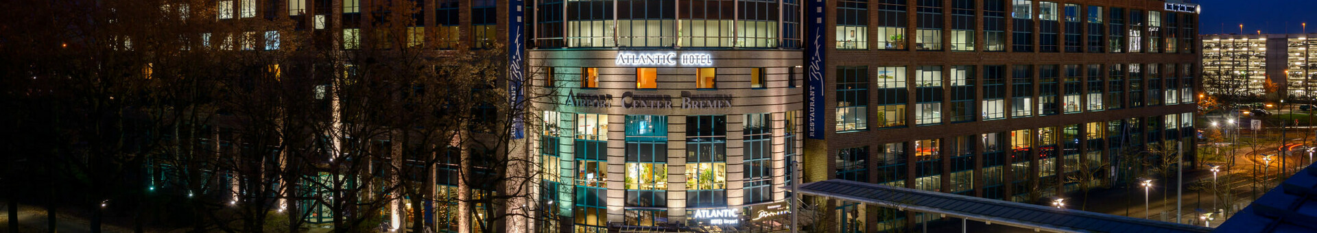 ATLANTIC Hotel Airport ATLANTIC Hotel Airport bei Nacht, beleuchtetes modernes Gebäude mit Glasfassade und angrenzender Straßenbahnhaltestelle.