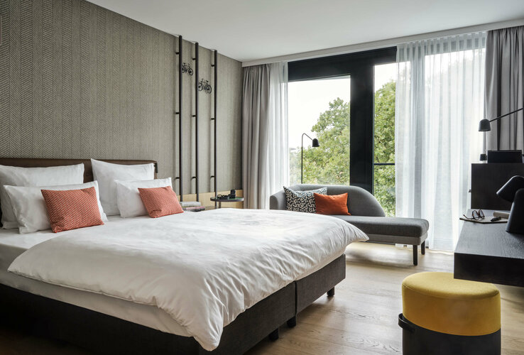 Comfort Zimmer in ATLANTIC Hotel Münster Modernes Hotelzimmer mit Doppelbett, Sofa, Schreibtisch und großen Fenstern mit Blick ins Grüne.