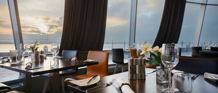 Gedeckte Tische im Restaurant STROM des ATLANTIC Hotel SAIL City Bremerhaven Modernes Restaurant mit gedeckten Tischen, großen Fenstern und Blick auf Sonnenuntergang über dem Wasser im ATLANTIC Hotel.