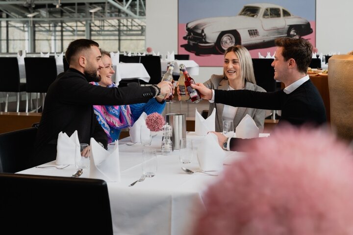 Gäste im Restaurant Gottlieb Bremen Vier Personen stoßen an einem elegant gedeckten Restauranttisch an, im Hintergrund ein Oldtimer-Bild.