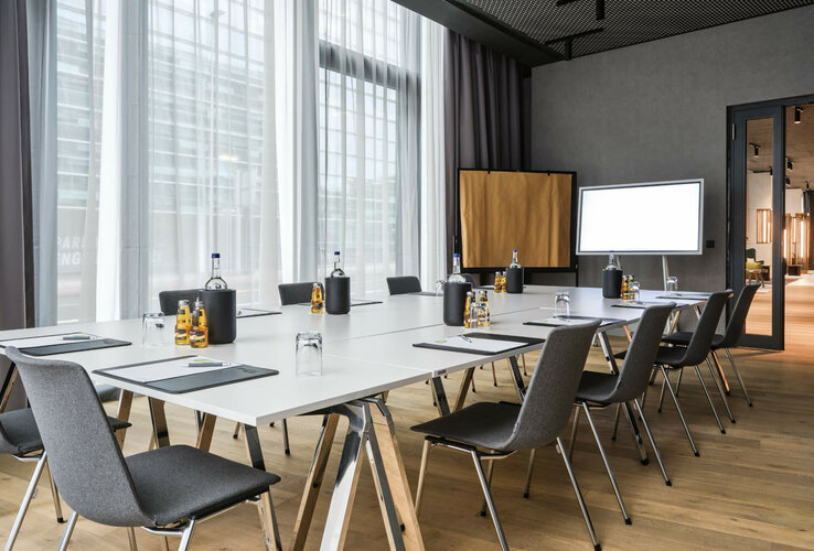 Tagen im ATLANTIC Hotel Münster Moderner Konferenzraum mit großem Tisch, Stühlen, Schreibmaterial, Wasserflaschen und natürlichem Licht im ATLANTIC Hotel Münster.