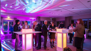 ATLANTIC Hotel Airport Bremen Events Feiern Tagen Eine Gruppe von Menschen steht an beleuchteten Stehtischen in einem farbig erleuchteten Veranstaltungsraum.