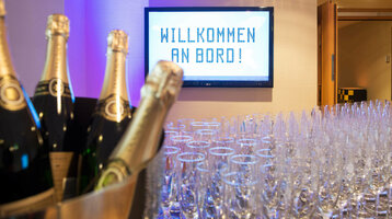 Sektempfang im ATLANTIC Hotel Airport Bremen Champagnerflaschen und Gläser vor einem Bildschirm mit "Willkommen an Bord!" im ATLANTIC Hotel Airport Bremen.