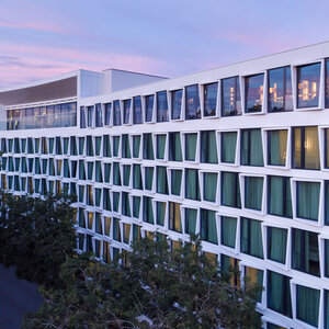 Aussenfassade ATLANTIC Hotel Münster Modernes Hotelgebäude mit großen Fenstern und grünen Vorhängen, umgeben von Bäumen, bei Sonnenuntergang.