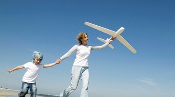 Mutter mit Kind spielen mit einem Flugzeug Eine Frau und ein Kind laufen am Strand, halten Hände und tragen weiße Kleidung, während sie ein Spielzeugflugzeug fliegen.