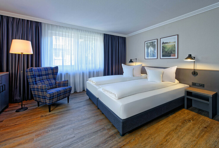 Zimmer im ATLANTIC Hotel Landgut Horn Bremen Helles Hotelzimmer mit Doppelbett, kariertem Sessel, Stehlampe, Holzboden und Bildern an der Wand.