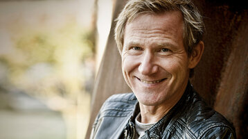 Krimi-Lesung im ATLANTIC Hotel Münster Eine Person lächelt in die Kamera, trägt eine schwarze Lederjacke, im Hintergrund unscharfe Natur.