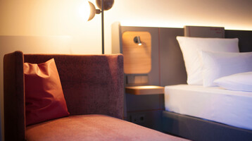 Hotelzimmer des ATLANTIC Hotel Airport in Bremen Modernes Hotelzimmer mit gemütlichem Sessel, Nachttischlampe und einem Bett mit weißen Kissen im ATLANTIC Hotel Airport.