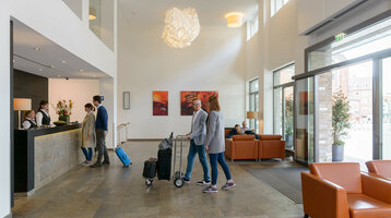 ATLANTIC Hotel Lübeck – Foyer Helle Hotel-Lobby mit Gästen an der Rezeption, modernen Möbeln und großen Fenstern im ATLANTIC Hotel Lübeck.