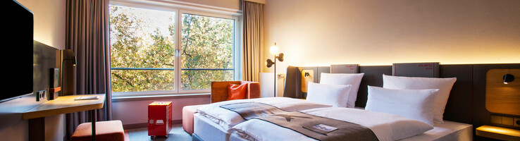 Business Zimmer im ATLANTIC Hotel Airport Modernes Hotelzimmer mit Doppelbett, Schreibtisch, großem Fenster und Decke mit Wolkenmotiv im ATLANTIC Hotel Airport Bremen.