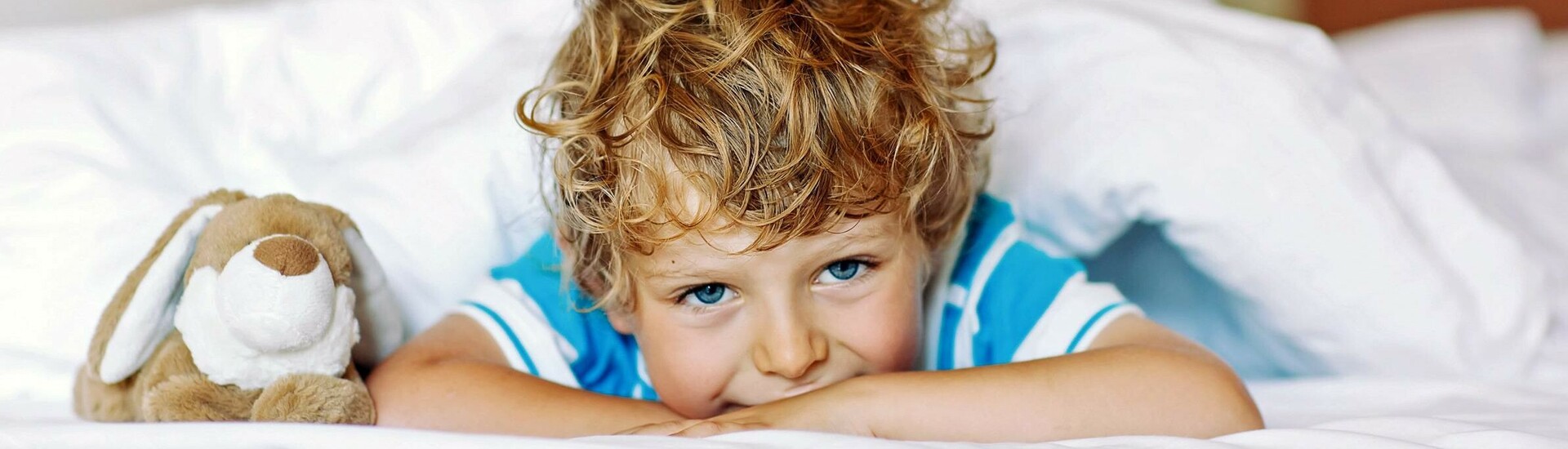 Kind mit Hase Kuscheltier im Bett der Familie Kind mit lockigem Haar liegt lächelnd neben Kuscheltier auf weißem Bett in einem Hotelzimmer.