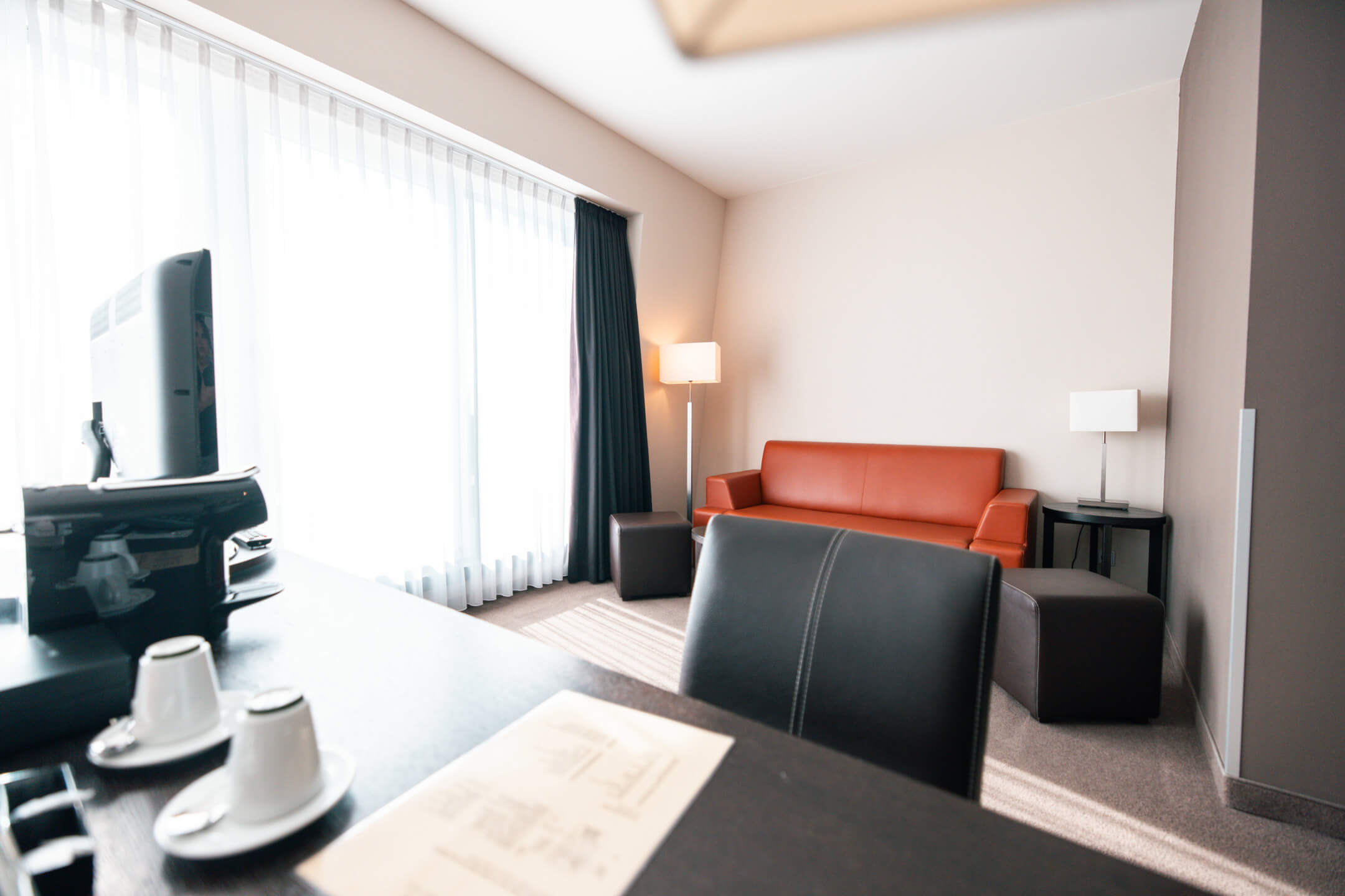 ATLANTIC Hotel Lübeck Studio Wohnbereich Bright hotel room with red sofa, desk, TV, two coffee cups and large windows with curtains.