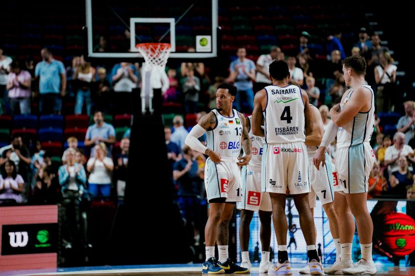 Basketball players in white jerseys stand on the court, spectators clap in the background.