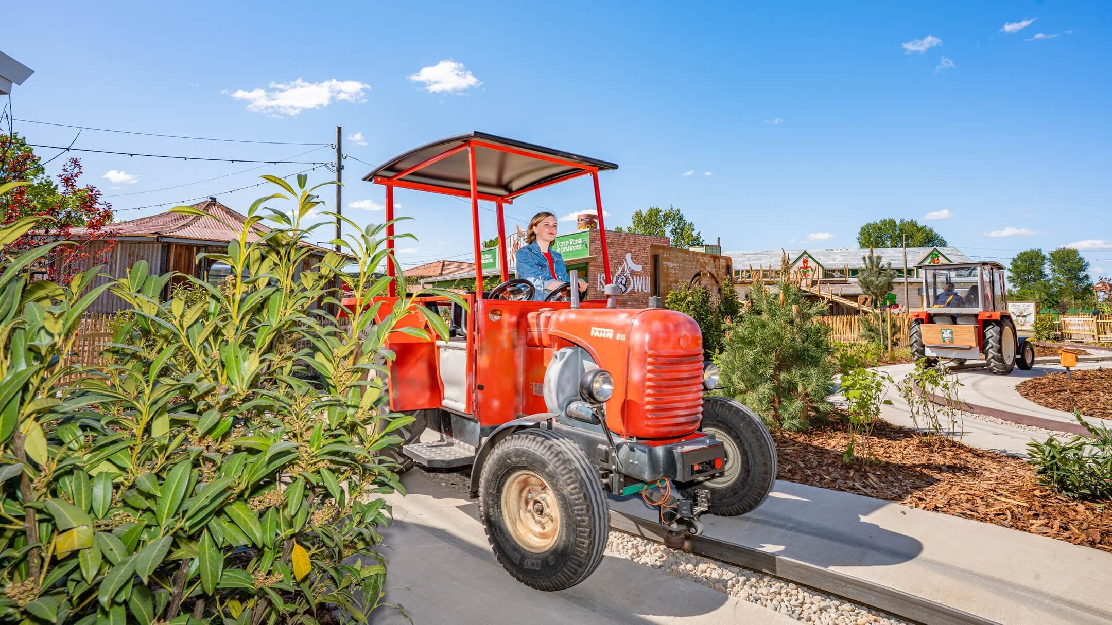 ATLANTIC Hotel Sail City Treggerbahn in Karls Erlebnis-Dorf Loxstedt Person driving red tractor on sunny hotel grounds with plants and wooden houses in the background.