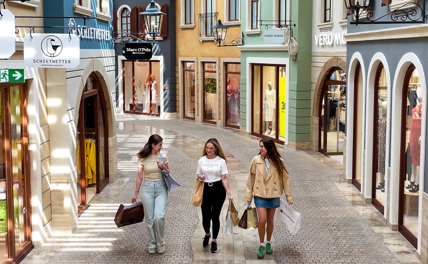 Three smiling women stroll through an elegant shopping street with boutiques.