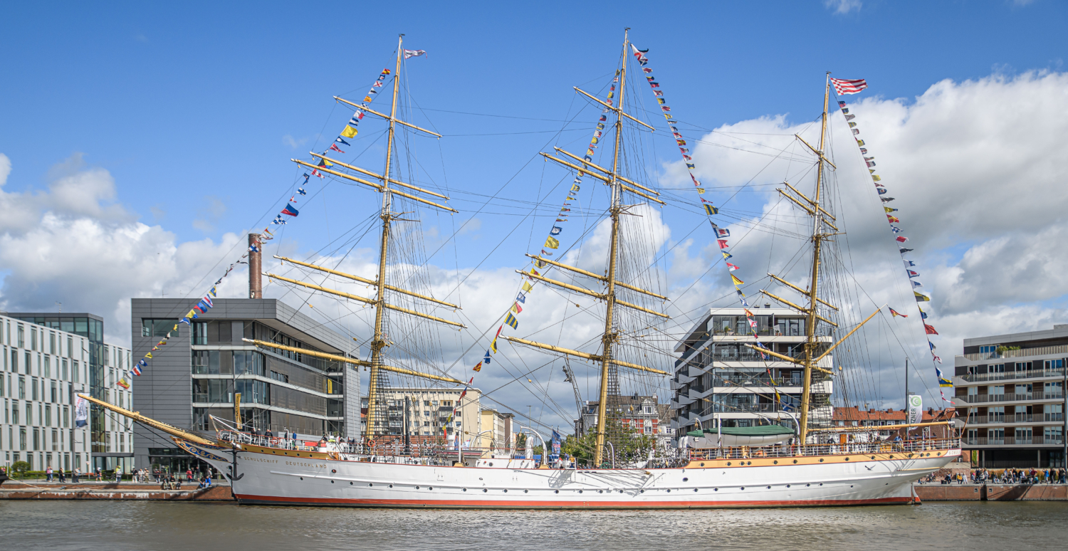 Large sailing ship with colorful flags, moored at a quay in front of modern buildings under a blue sky.